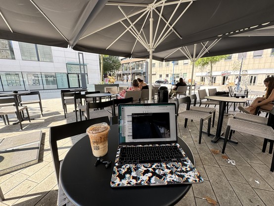 Outdoor café with tables and chairs under large umbrellas. A laptop and iced coffee are on one table. People are sitting and relaxing in the background.
