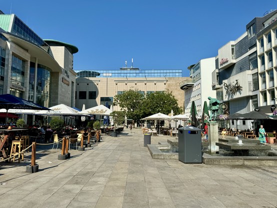 A vibrant city square featuring outdoor cafes with white and black umbrellas, a statue in the center, and modern buildings in the background under a clear blue sky.