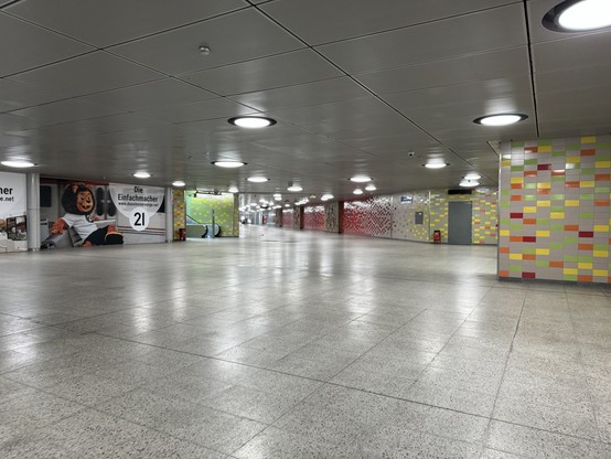 An empty, well-lit underground walkway with tiled walls in various colors, illuminated ceiling lights, and a large advertisement featuring a character on the left wall.