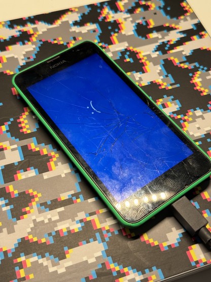 A cracked-screen Nokia Windows Phone with a BSOD, placed on a colorful pixelated surface, connected to a charging cable.