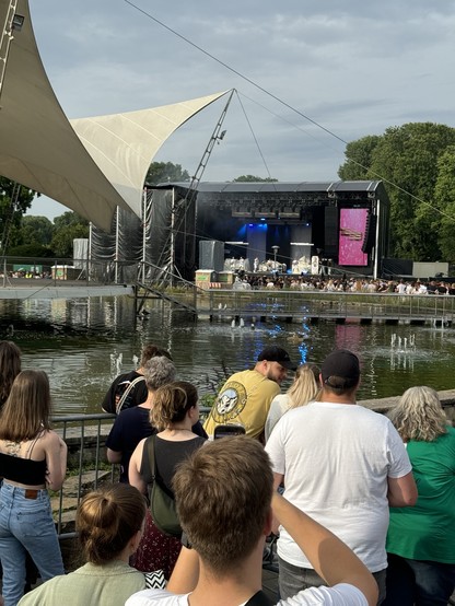 Audience watching a live outdoor concert by a water feature, with a large stage setup and speakers, surrounded by trees and under a partly cloudy sky.