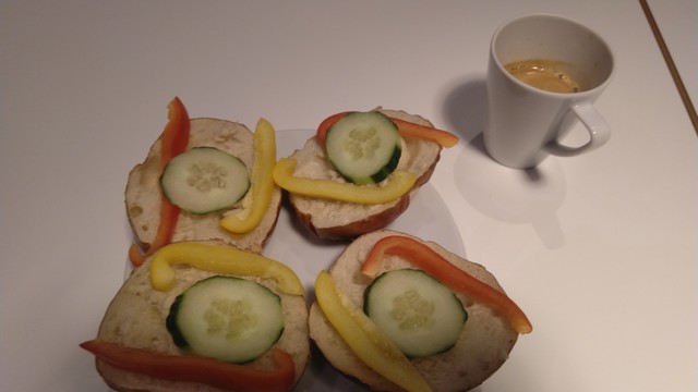 Bild eines Hotelfrühstücks.
Auf einem Teller liegen zwei aufgeschnittene Brötchen. Auf den trockenen vier Hälften liegen jeweils eine Scheibe Gurke sowie ein roter und ein gelber Paprikastreifen. Daneben steht ein doppelter Espresso