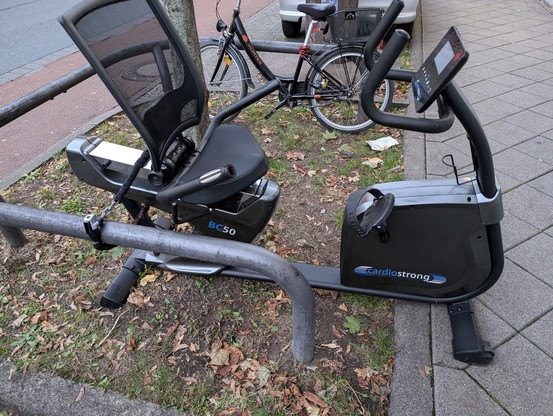 Ein Heimtrainer-Sitz-Fahrrad steht draußen neben dem Gehsteig, angekettet mit einem Fahrradschloss