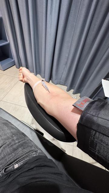 A close-up of a bare arm with a syringe inserted, presumably for a medical procedure. The background features gray curtains and a dark chair. A band is seen around the arm, and part of a person's leg is visible in the lower left corner.